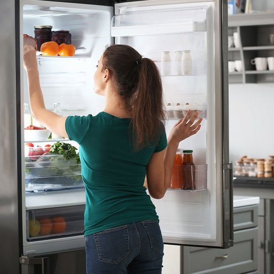 Eine Frau steht vor dem offenen Kühlschrank und greift nach etwas im obersten Fach