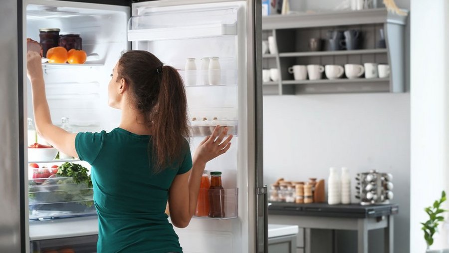 Eine Frau steht vor dem offenen Kühlschrank und greift nach etwas im obersten Fach